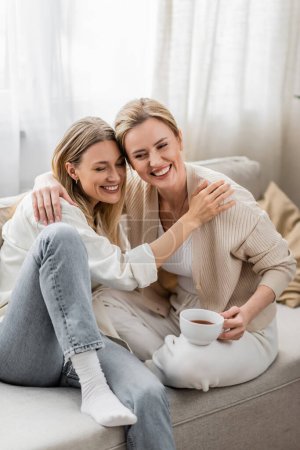 Photo for Two good looking pretty sisters in trendy attire sitting on sofa and hugging, family bonding - Royalty Free Image