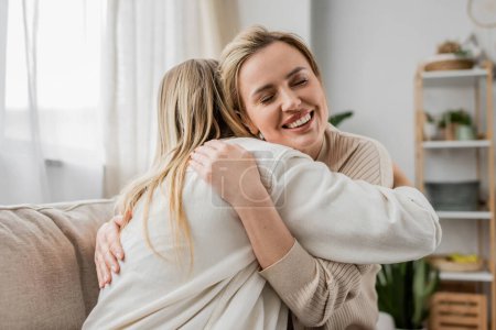 Photo for Two blonde sisters in lovely casual attire hugging on sofa with closed eyes, family bonding - Royalty Free Image
