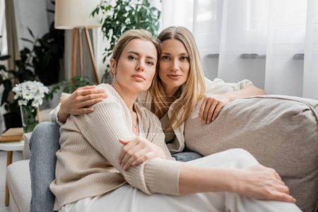 Porträt hübscher Schwestern, die auf dem Sofa sitzen, sich umarmen und in die Kamera schauen, familiäre Bindung