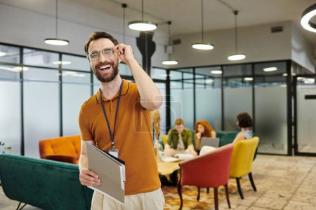Téléchargez les photos : Entrepreneur barbu réussie dans les lunettes souriant à la caméra près de l'équipe parlant dans le bureau moderne - en image libre de droit