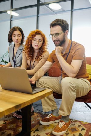 Teamwork, lächelnder bärtiger Unternehmer, der neben lächelnden Geschäftsfrauen im modernen Büro auf den Laptop schaut