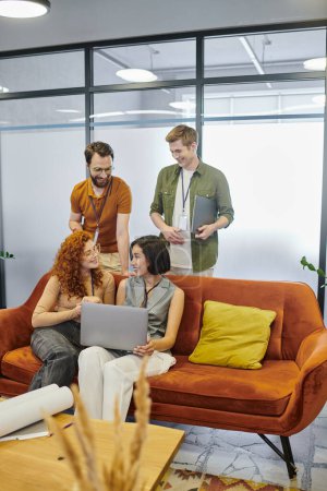 positive Geschäftsleute in der Nähe junger Frauen mit Laptop im Gespräch auf der Couch im modernen Büro, Startup-Projekt