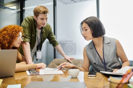 jeune gestionnaire pointant vers des graphiques près de rousse collègue et chef d'équipe au bureau, projet d'entreprise