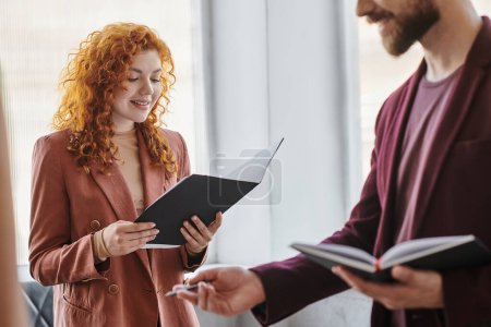 positive redhead businesswoman looking at documents near colleague in coworking office