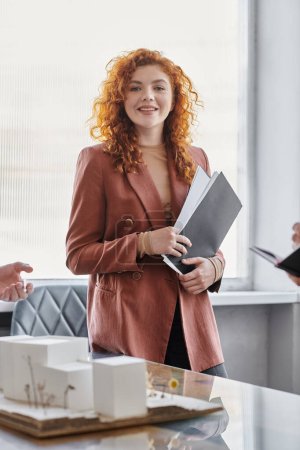 portrait of redhead architect with documents smiling at camera near modern building model in office