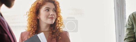 positive redhead businesswoman with documents looking away near colleague in office, banner
