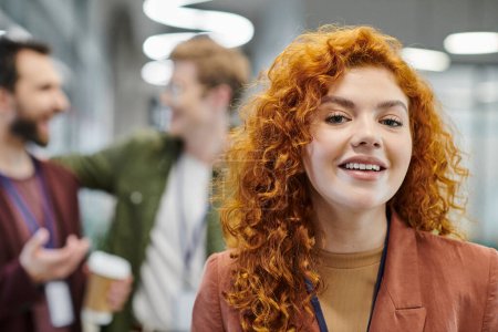portrait of optimistic redhead businesswoman looking at camera near blurred colleagues in office