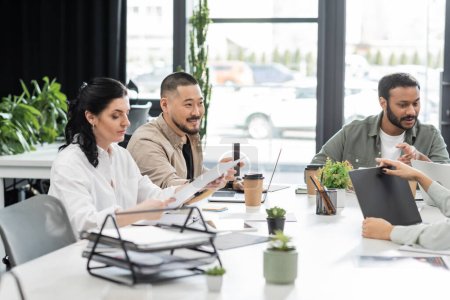 happy asian man showing smartphone with blank screen near multiethnic colleagues in modern coworking
