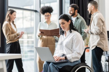 mujer de negocios discapacitada en silla de ruedas usando portátil cerca de colegas multiétnicos borrosos, inclusión