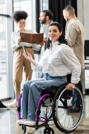 Glückliche behinderte Geschäftsfrau im Rollstuhl blickt in die Kamera neben verschwommenen multiethnischen Kollegen