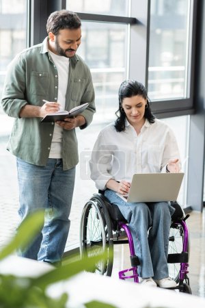 hombre indio feliz tomando notas cerca de la mujer de negocios discapacitada en silla de ruedas utilizando el ordenador portátil en la oficina