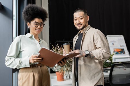 asian businessman with coffee and african american businesswoman looking at camera in office