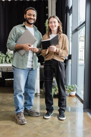 cheerful woman with notebook and indian man with laptop looking at camera in office, coworking space