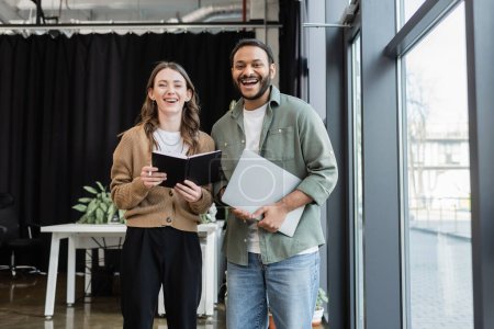 joyful woman with notebook and indian man with laptop looking at camera in office, coworking space