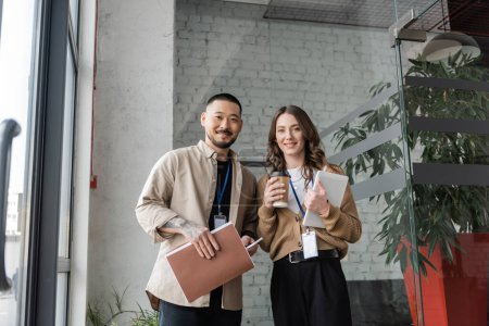 happy woman holding paper cup with coffee and tablet near asian businessman during break time