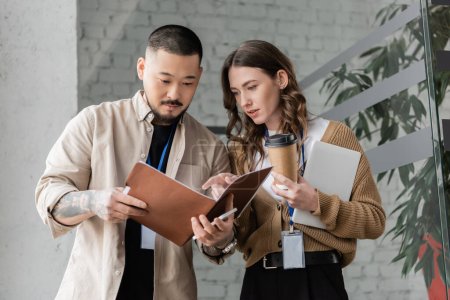 pretty businesswoman pointing at folder and discussing startup project with focused asian colleague