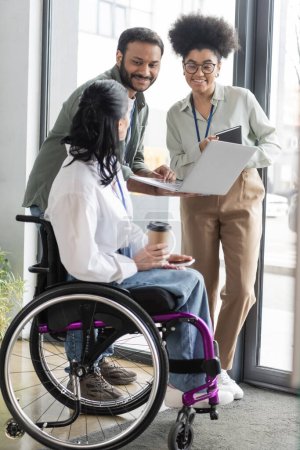 happy interracial colleagues looking at disabled coworker on wheelchair, discussing work strategy