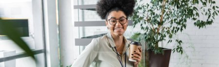 happy african american businesswoman in glasses holding paper cup with coffee in office, banner