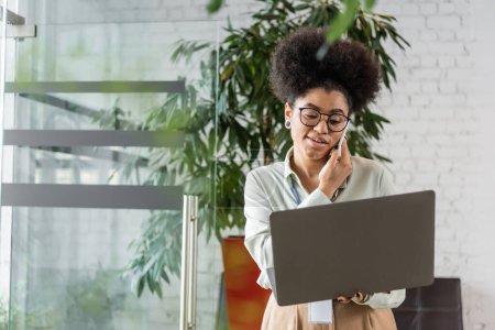 femme d'affaires afro-américaine dans des lunettes à l'aide d'un ordinateur portable et parler sur smartphone, multitâche