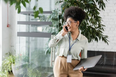 happy african american businesswoman in glasses holding laptop and talking on smartphone in office