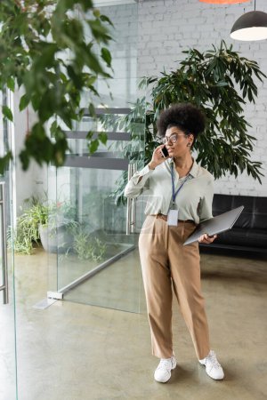 african american businesswoman in glasses holding laptop and talking on smartphone, full length