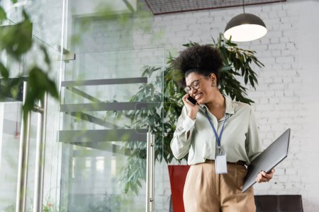 joyful african american businesswoman in glasses holding laptop and talking on smartphone in office