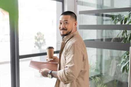 complacido asiático hombre de negocios con tatuaje sonriendo y caminando en oficina con carpeta y papel taza