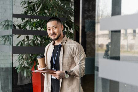 Foto de Feliz asiático hombre de negocios con tatuaje sonriendo y de pie con carpeta y café en papel taza - Imagen libre de derechos