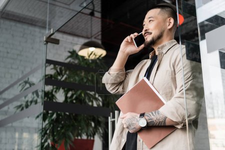 happy asian businessman with tattoo holding folder and talking on smartphone near glass door