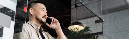 glücklicher asiatischer Mann mit Geschäftstelefon auf Smartphone in der Nähe von Glastür im Büro, Banner