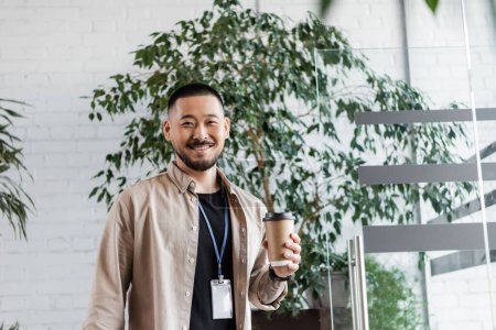 glücklicher asiatischer Geschäftsmann mit Tätowierung, der Coffee to go hält und in die Kamera schaut, Firmenleben