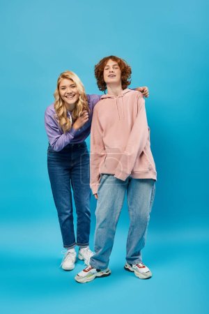 Foto de Alegre y elegante adolescente amigos en sudaderas con capucha y jeans sonriendo a la cámara en azul, longitud completa - Imagen libre de derechos