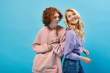 Photo for Excited blonde teen girl laughing at camera near smiling redhead boyfriend on blue - Royalty Free Image