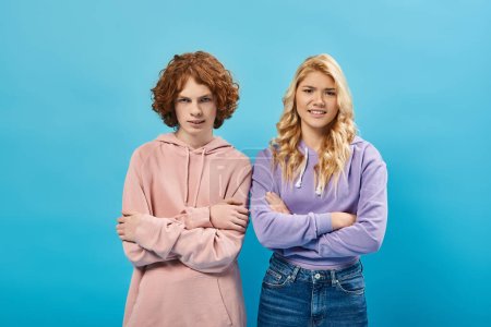 Photo for Irritated teenage friends in hoodies standing with crossed arms and looking at camera on blue - Royalty Free Image