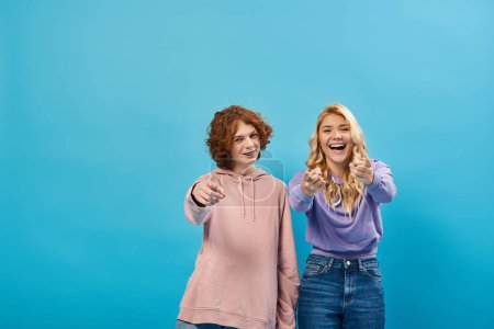 fröhliche und trendige Teenager-Freunde in Kapuzenpullis, die mit den Fingern in die Kamera zeigen und über blau lachen