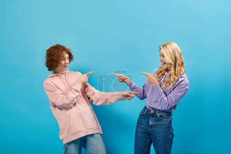joyful teenagers in stylish outfits pointing with fingers and each other and laughing on blue