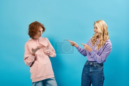 blonde teenage girl laughing and pointing with fingers at discouraged redhead boyfriend on blue