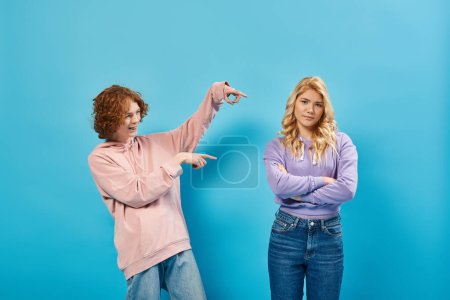 lachender rothaariger Teenie-Typ zeigt auf beleidigte Freundin, die mit verschränkten Armen auf blauem Grund steht