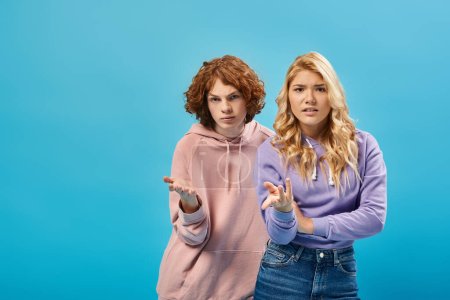 Photo for Displeased teenage boy and girl in stylish hoodies pointing with hands and looking at camera on blue - Royalty Free Image
