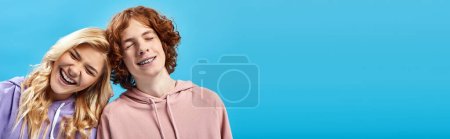 Photo for Portrait of cheerful teenage guy and girl in hoodies laughing with closed eyes on blue, banner - Royalty Free Image