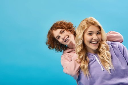cheerful redhead teen boy with blonde girlfriend having fun and laughing at camera on blue