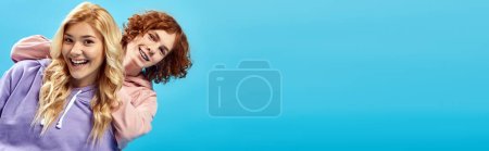 blonde teenage girl with redhead having fun in studio and laughing at camera on blue, banner