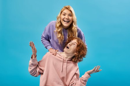 Photo for Excited blonde teen girl having fun and laughing at camera near surprised redhead boyfriend on blue - Royalty Free Image