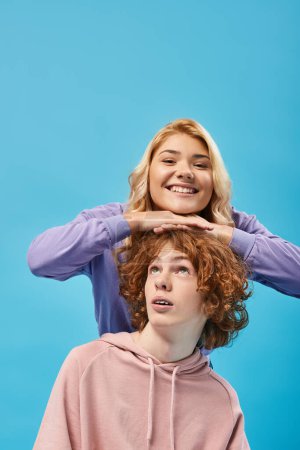 expressive blonde teen girl having fun and smiling at camera above head of redhead boyfriend on blue