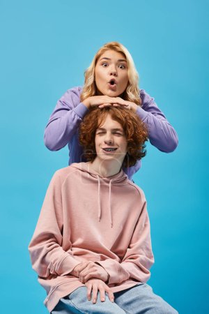 Adolescente blonde étonnée avec bouche ouverte regardant la caméra au-dessus de la tête du petit ami rousse sur bleu