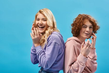 Photo for Excited teenage friends in hoodies sitting back to back and laughing at camera in studio on blue - Royalty Free Image