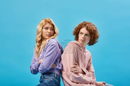 Photo for Offended teenage friends with folded arms sitting back to back and looking at each other on blue - Royalty Free Image