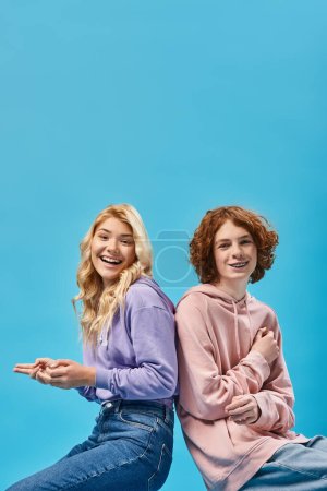 fröhliche Teenager-Freunde in trendigen Kapuzenpullovern, die Rücken an Rücken lachend in die Kamera im Studio auf blauem Grund sitzen