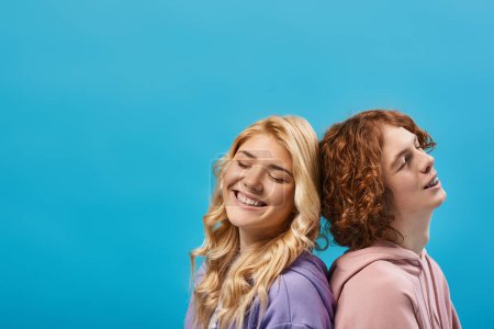 couple adolescent positif en sweat à capuche élégant assis dos à dos et souriant avec les yeux fermés sur le bleu