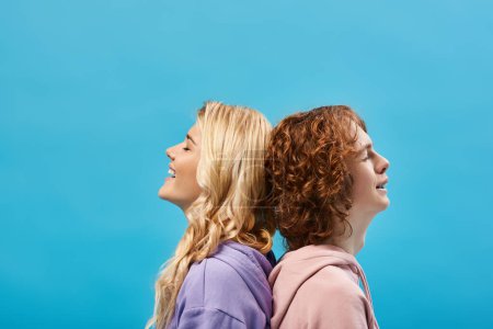 Photo for Carefree teenage friends sitting back to back and smiling with closed eyes on blue, side view - Royalty Free Image